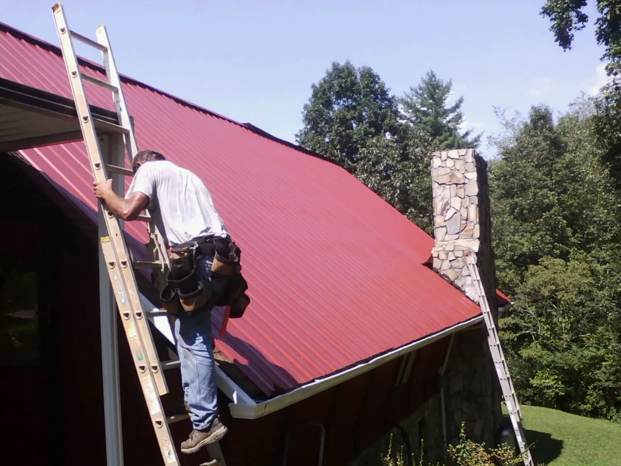 Roofer on a ladder beside a red metal roof and stone chimney — Soffit & Fascia Repair by Roofing Brothers in Port Ludlow
